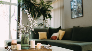 A cozy living room with a green sofa, cushions, a wooden tray with candles, and a vase of blooming branches.