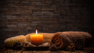 Glowing candle surrounded by spa items, towels, and pebbles, against a textured stone wall.