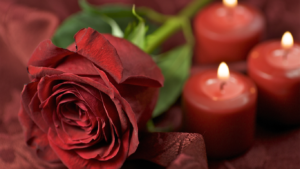 Close-up of a red rose and three glowing candles on a dark red fabric background, creating romance.